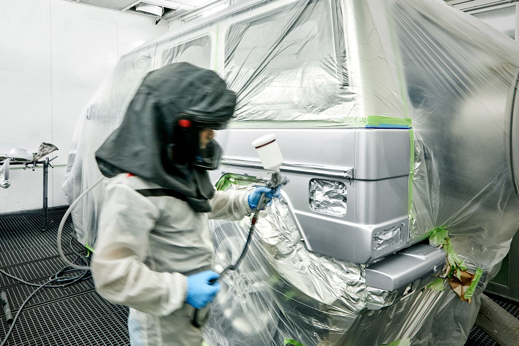 Lackierer in Schutzanzug lackiert ein abgeklebtes Nutzfahrzeug in der Lackierkabine